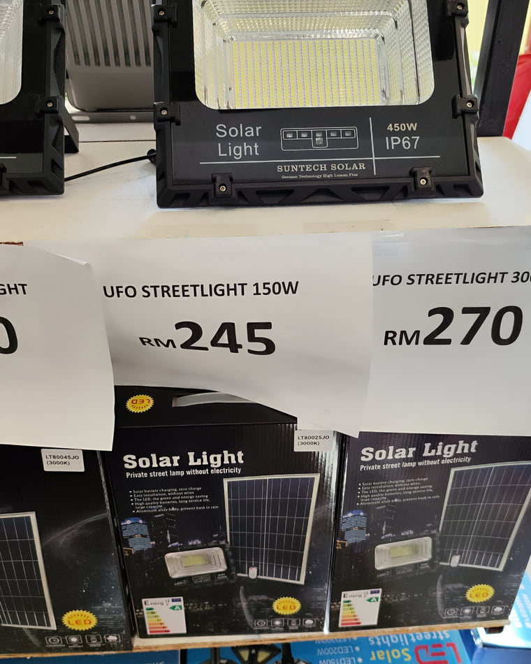 Two large solar street-light heads on a counter above printed shop price cards.
